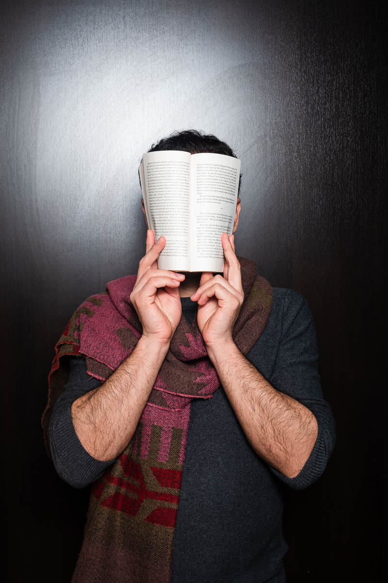 Portrait of refugee Dewran hiding his face with an open book facing the camera
