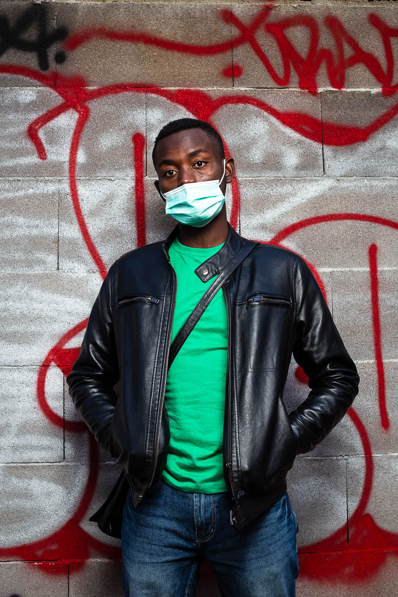 Portrait of refugee Franky wearing a face mask with his hands in the pockets of his black leather jacket against a graffiti wall