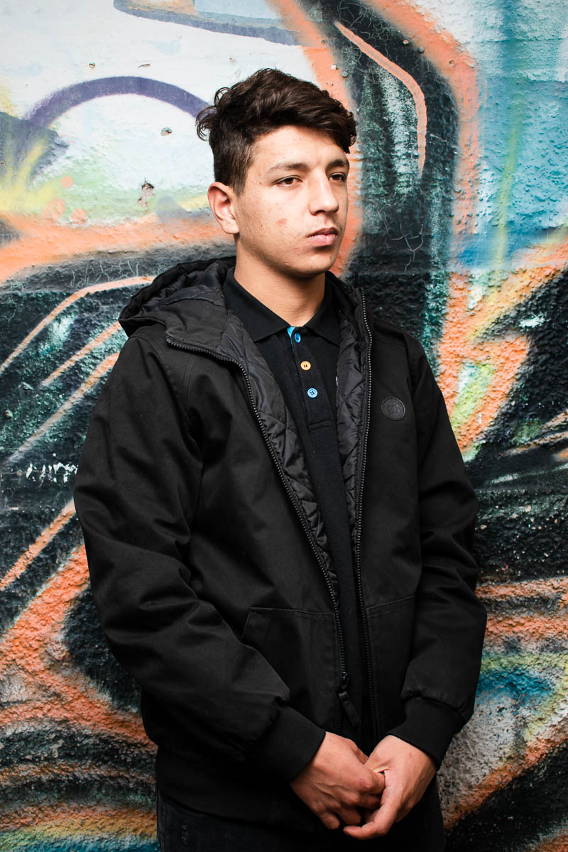 Portrait of refugee Mhieadeen wearing a black jacket with his hands clasped looking to his left standing against a graffiti wall
