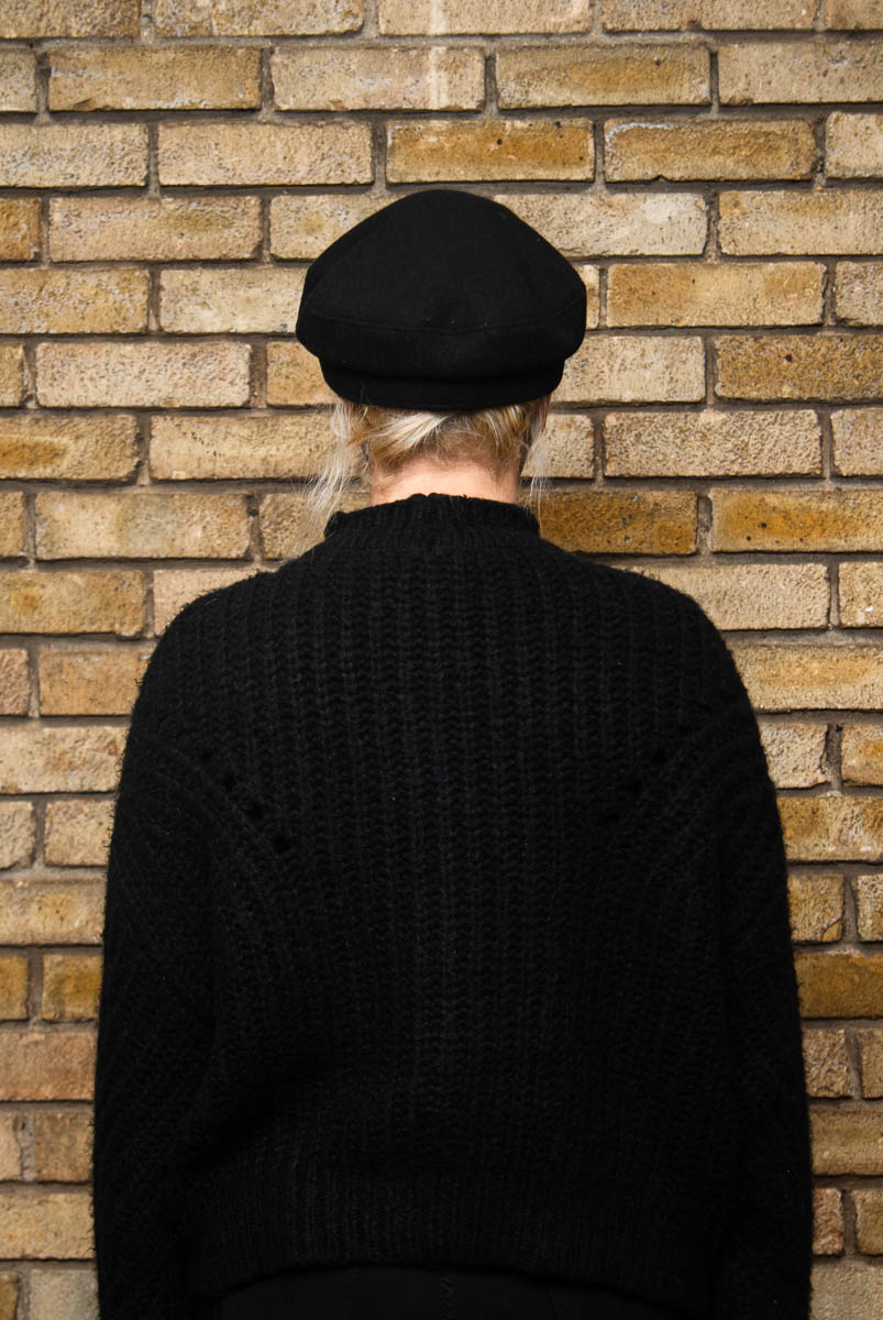 Portrait of refugee Sara with her back facing the camera wearing a black knitted sweater with a beret turned towards a brick wall