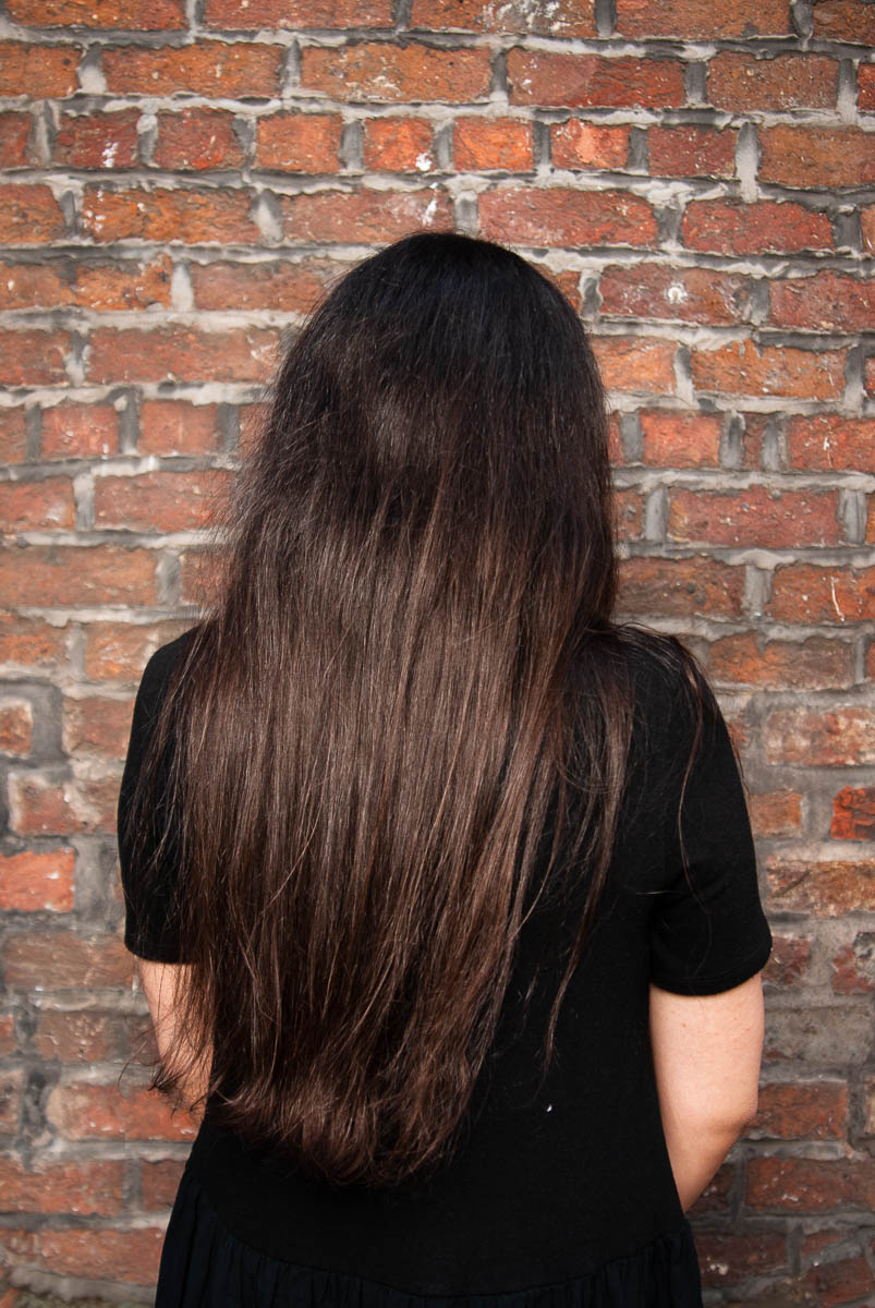 Portrait of refugee Marie with her back completely facing the camera against a brick wall