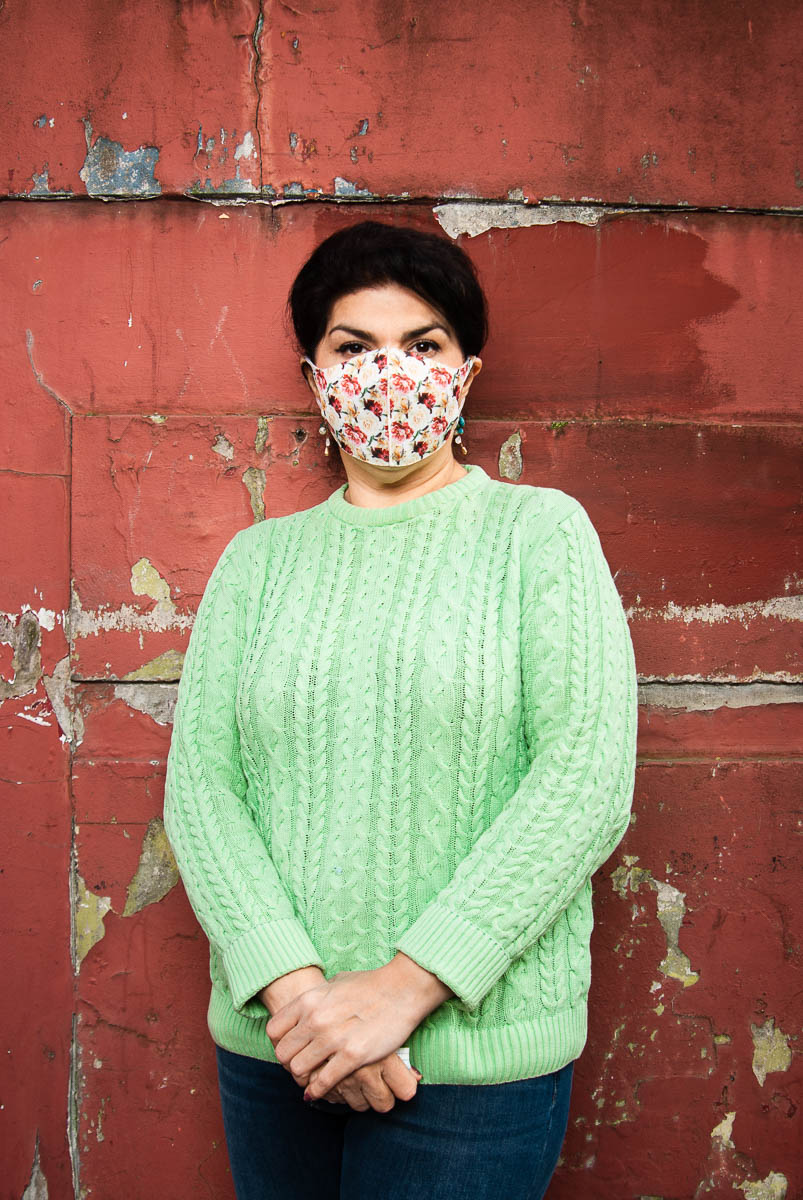 Portrait of refugee Maryam wearing a lime green knitted sweater and a face mask with her hands folded standing against a old wall