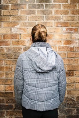 Portrait of refugee Shima wearing a checkered hoodie jacket turned away facing the brick wall with their hair in a bun