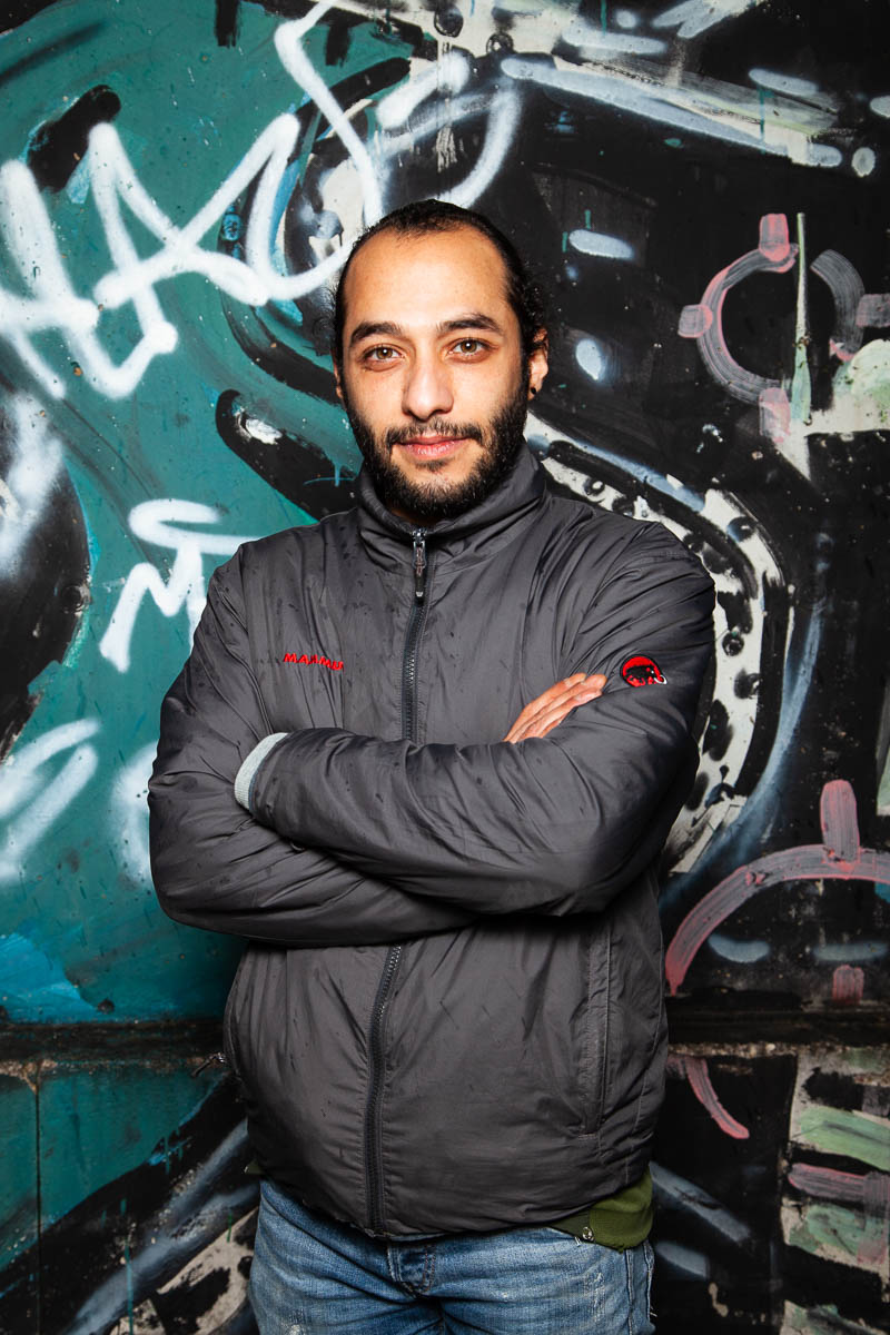 Portrait of refugee Mohamad wearing a gray jacket with his arms crossed standing against a graffiti wall