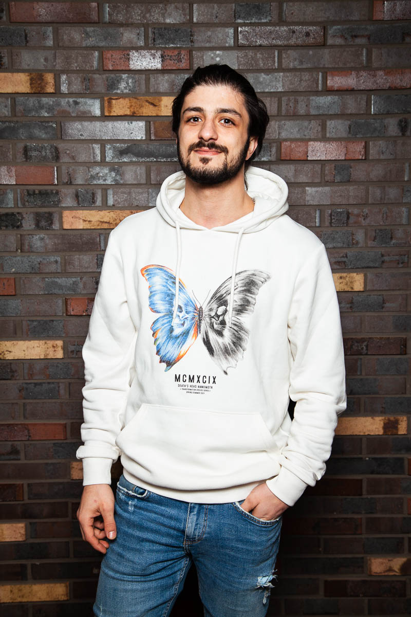 Portrait of refugee Jihad wearing a white hoodie with a printed butterfly standing with his hand in the pocket of his jeans standing against a brick wall
