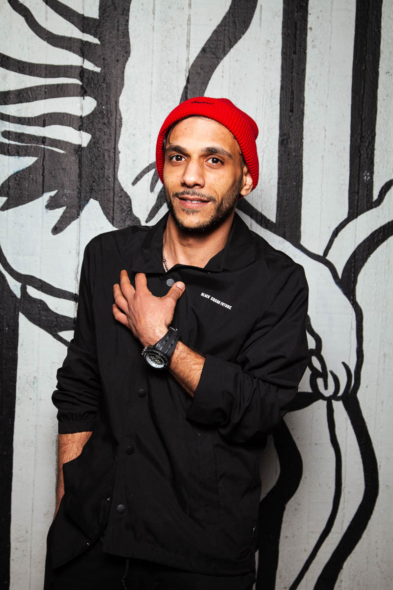Portrait of refugee Wael with his left hand on his chest wearing a red beanie standing against a graffiti wall