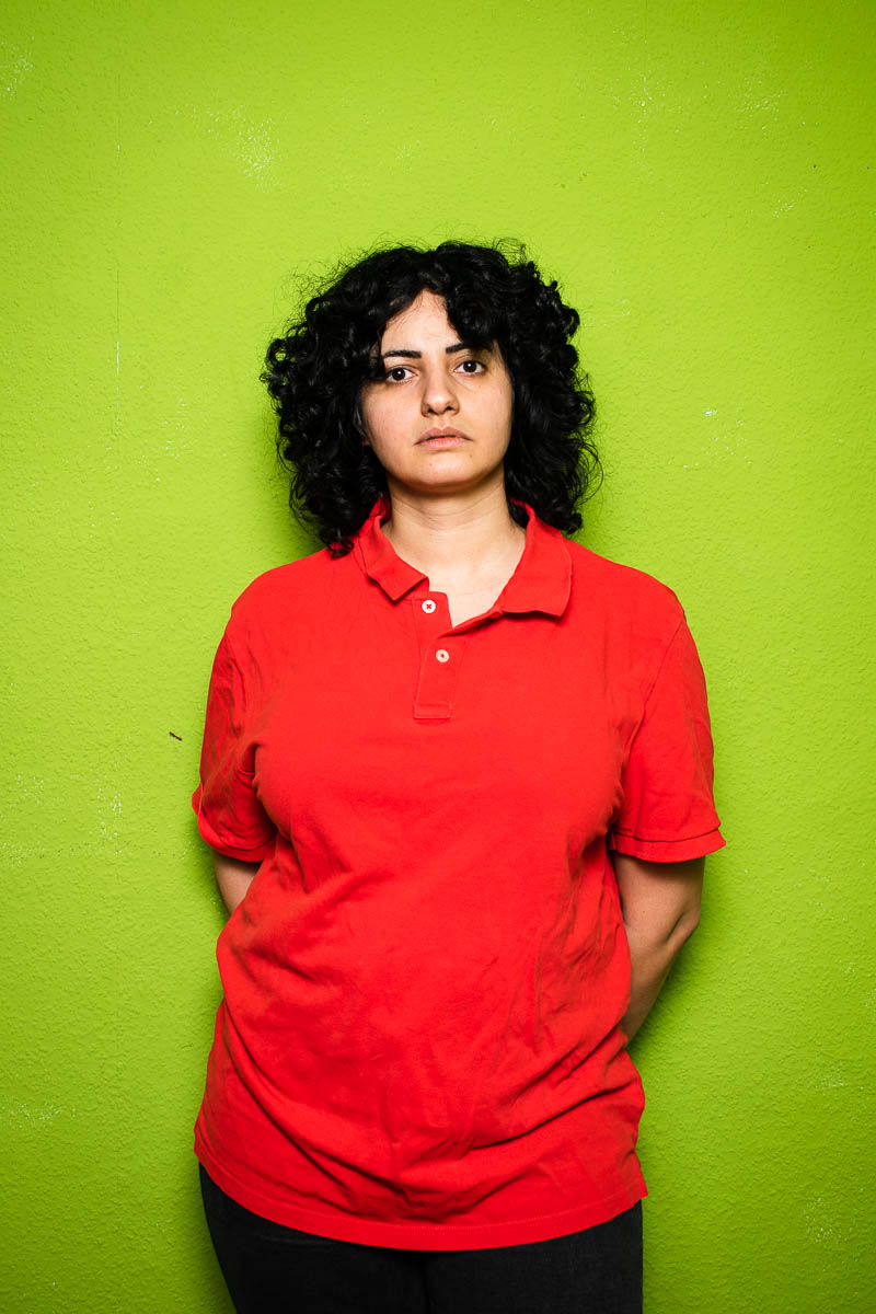 Portrait of refugee Yasaman wearing a red polo shirt with hands behind the back standing against a green wall