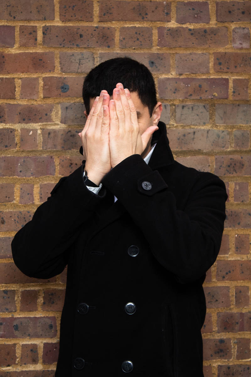 Portrait of refugee Remy hiding his face with both his hands standing against a brick wall