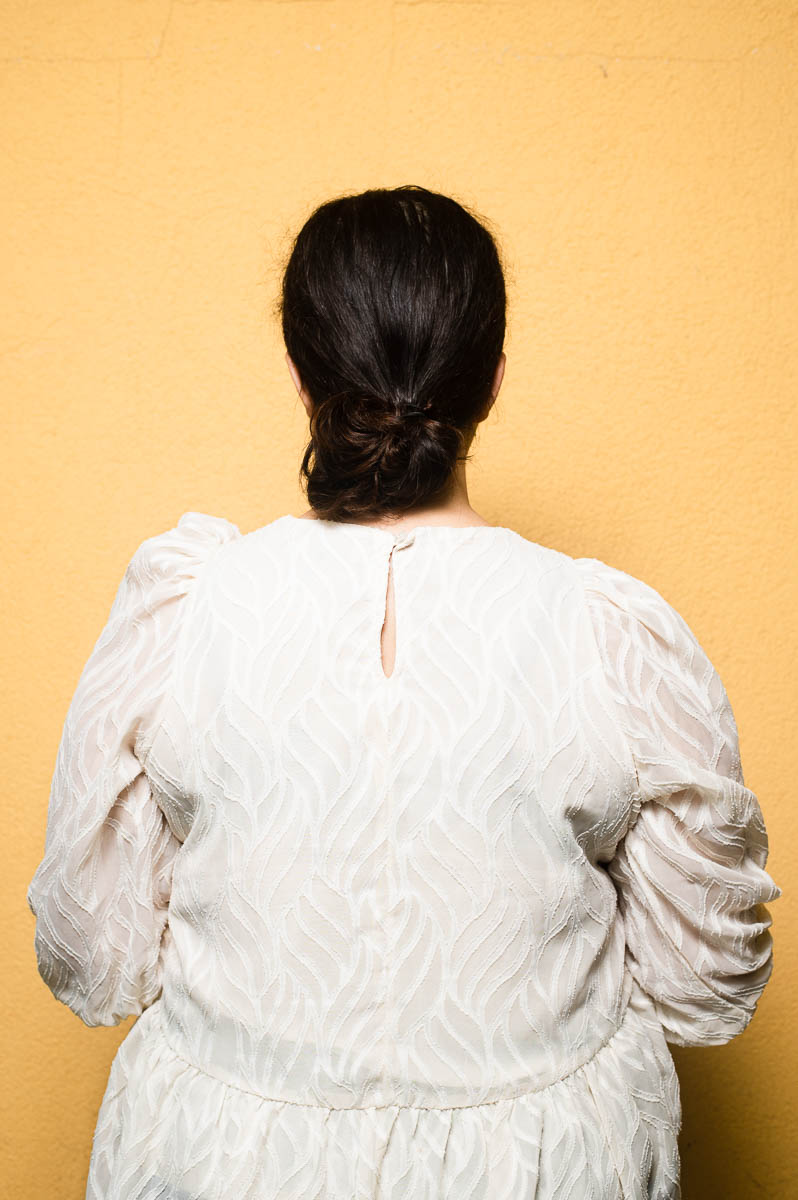 Portrait of refugee Jasmin wearing a white lace dress with her back completely turned away and facing the camera