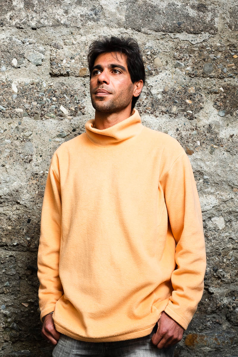 Portrait of refugee Mo wearing a orange turtleneck with his hands in his pockets standing against a stone wall