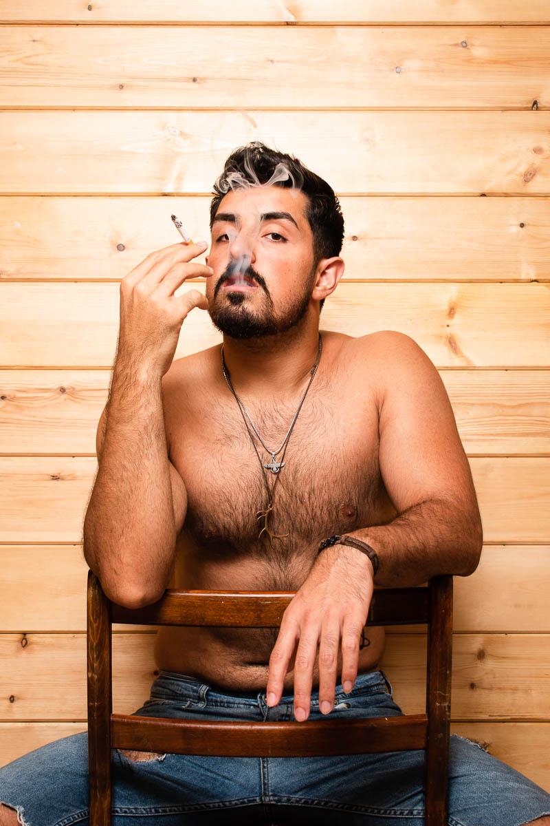 Portrait of refugee Shiar shirtless sitting on a flipped wooden chair with one hand on the top of the chair and the other holding a cigarette he is smoking