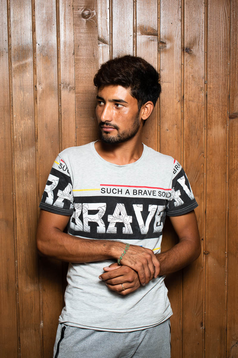 Portrait of refugee Hanif with his right hand holding his left wrist looking to his right and standing against a wooden planked wall