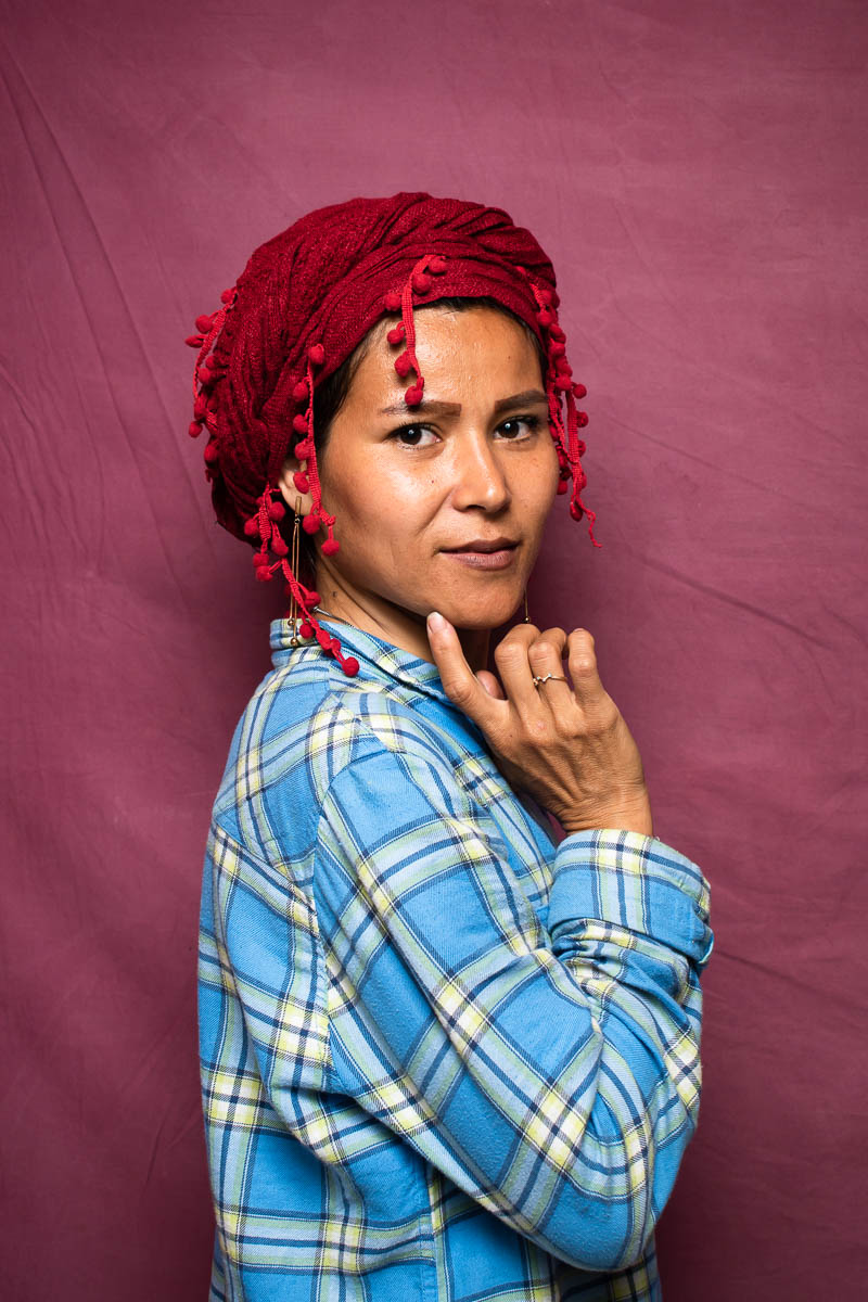 Portrait of refugee Fateme wearing a red hijab turban standing sideways with her right hand and forefinger pointed to her chin