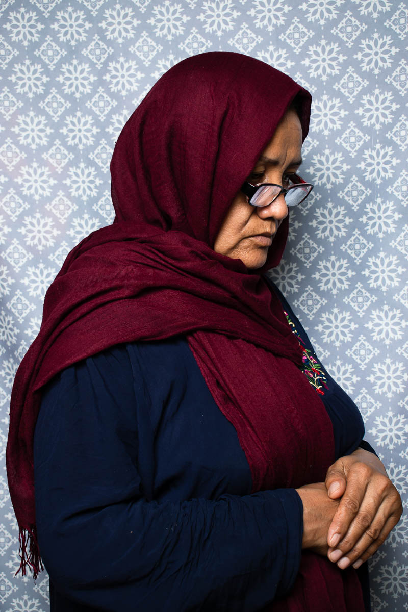 Portrait of refugee Sakine standing sideways wearing a maroon hijab and glasses looking down to her hands folded and resting on her stomach