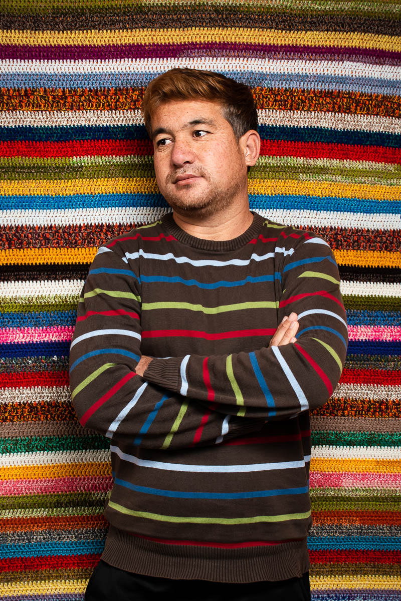 Portrait of refugee Sajad wearing a colored striped shirt with his arms crossed and looking to his right standing against a colorful woven striped tapestry