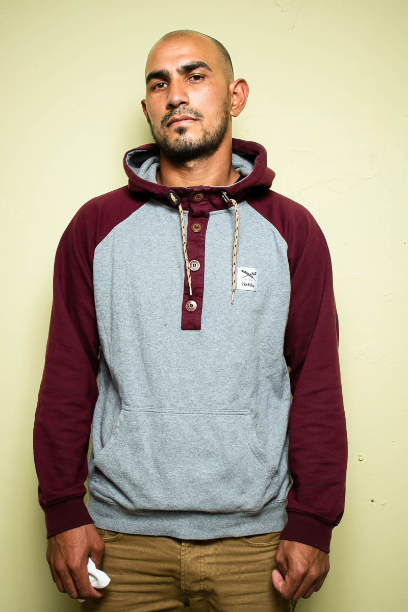 Portrait of refugee Qurban wearing a maroon and gray collared jacket with a white cloth crumpled in his right hand
