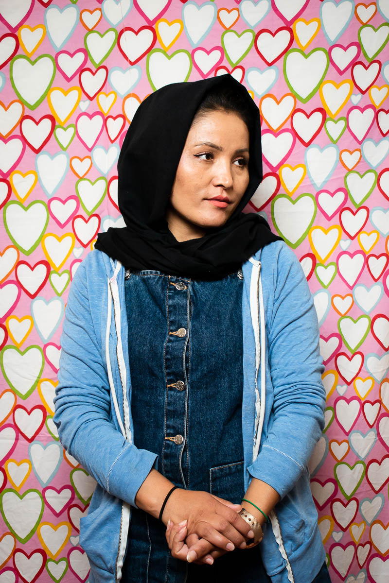 Portrait of refugee Latife wearing a black hijab looking to her left with her hands clasped standing against a background covered with colorful hearts