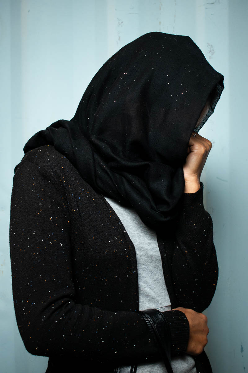 Portrait of refugee Narges turned to her left with hwe left hand fisted at her face and the right hand wrapped around ger stomach and her black hijab covering all her face