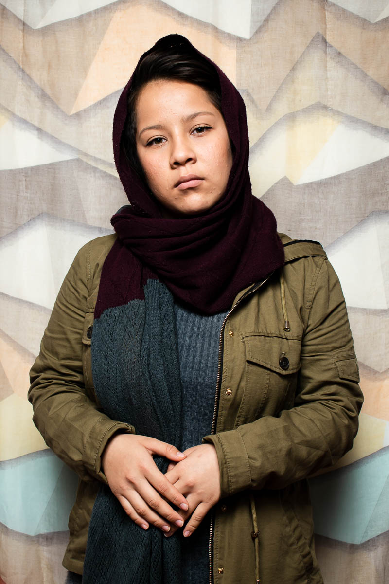 Portrait of refugee Fateme wearing a black hijab and dark green jacket with her hands folded and resting on her stomach