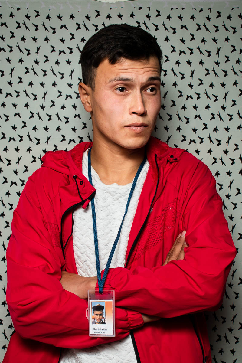 Portrait of refugee Ramin wearing a red jacket and a ID card with his hands crossed and looking to his left