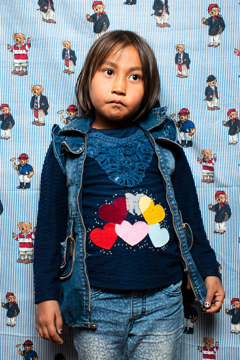 Portrait of a child refugee Nazanin wearing a denim jacket standing against a child's printed blanket