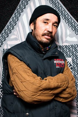 Portrait of refugee Mosa wearing a black cap and sleeveless puffer jacket over a brown sweater with his hands crossed