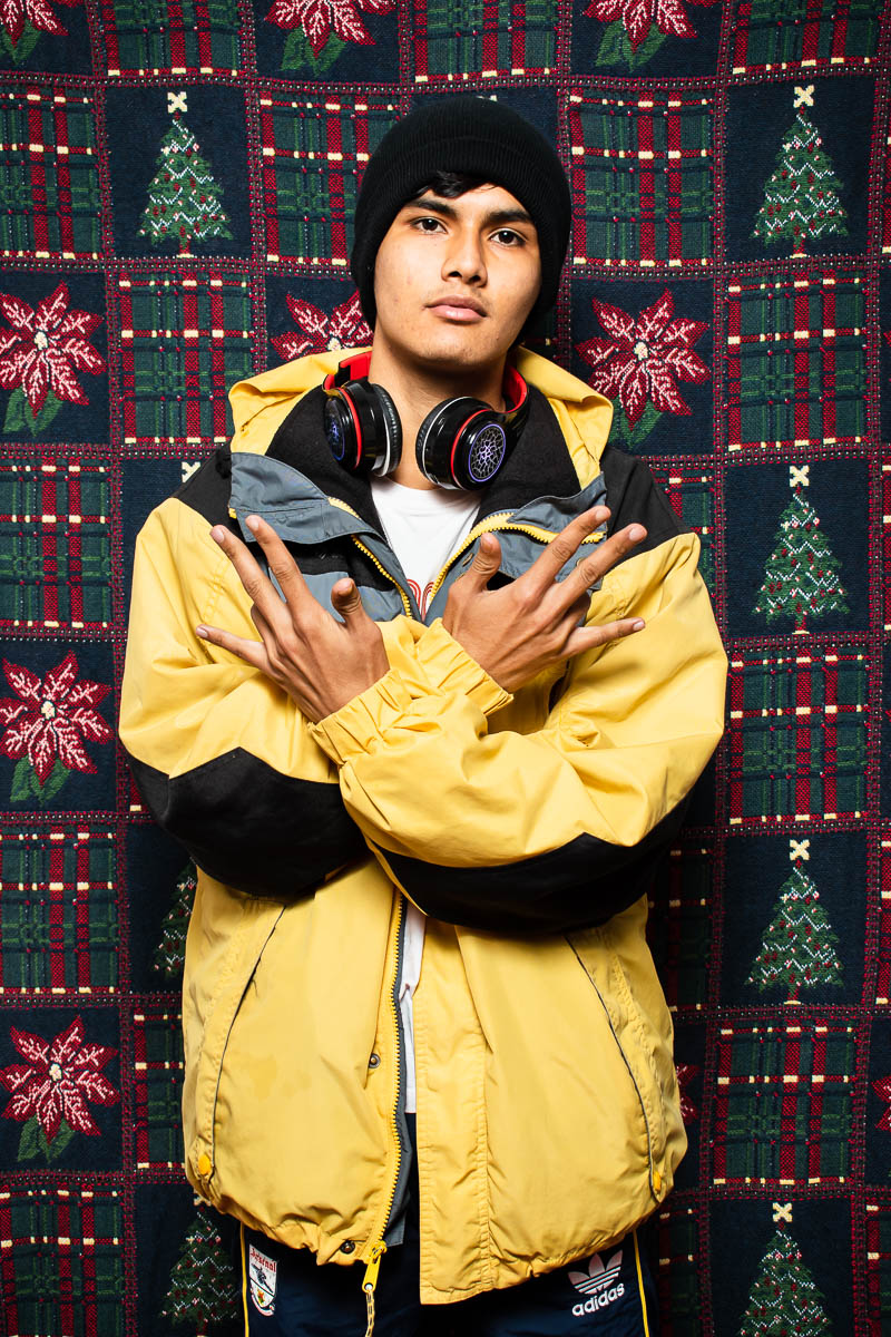 Portrait of refugee Amir wearing a yellow jacket and a headset around his neck with both his hands raised in a gesture with only his ring fingers folded