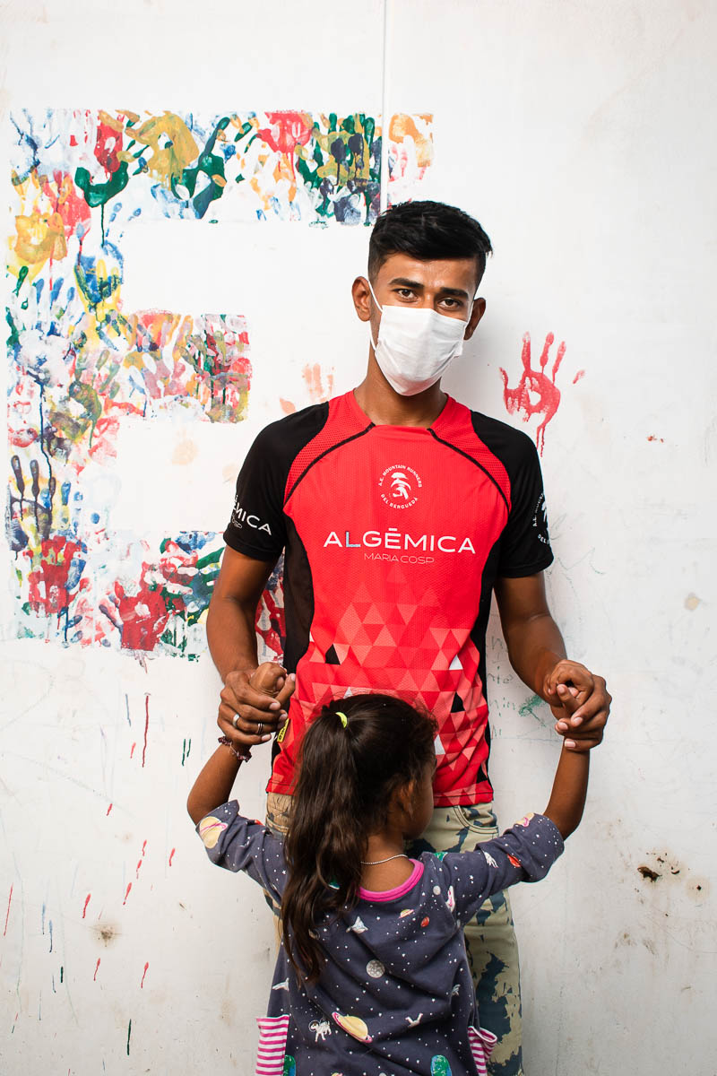 Portrait of refugee Alireza wearing a face mask and holding the hands of a little girl who is turned away from the camera facing him against a colorful handprint background