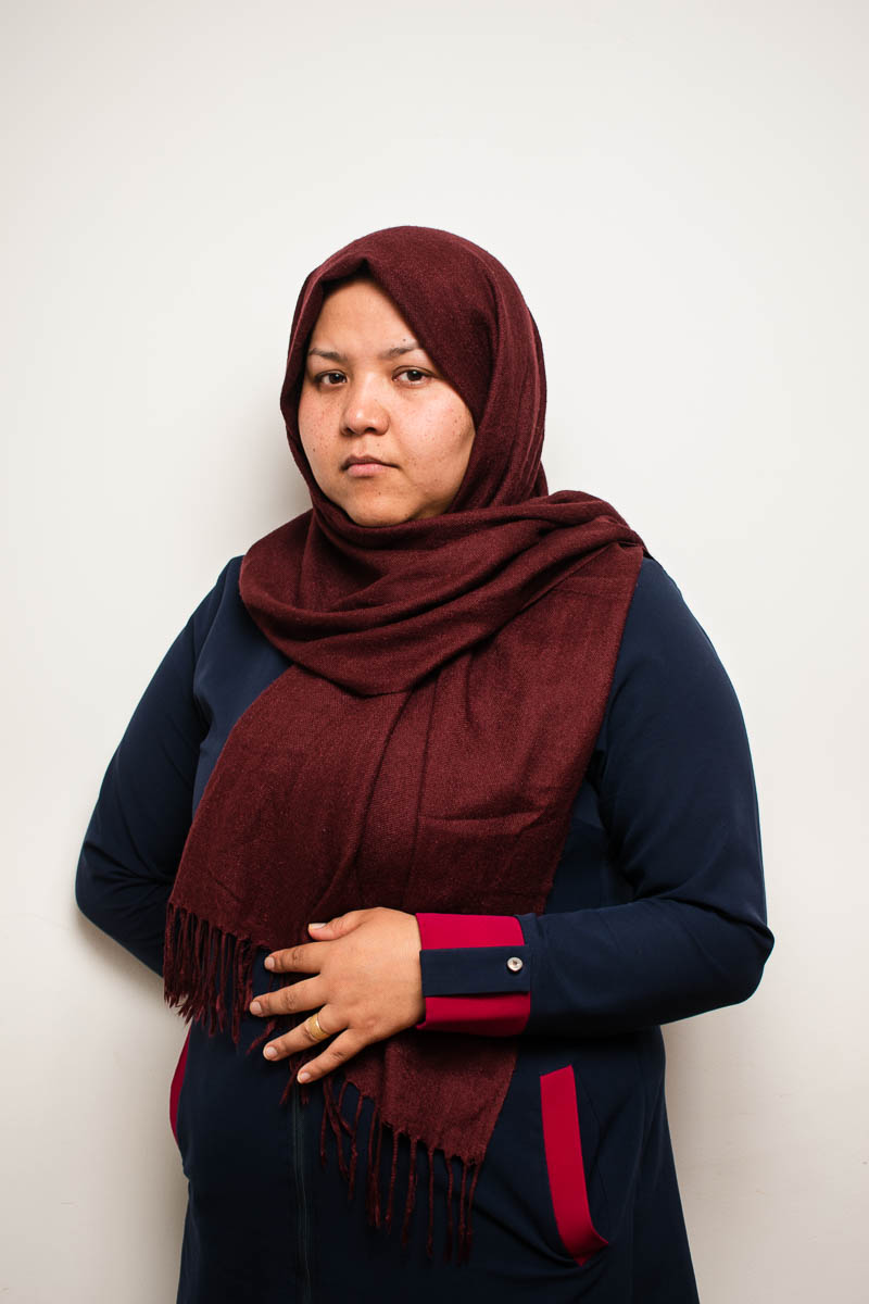 Portrait of refugee Aziza wearing a maroon hijab with one hand resting on her stomach and the other behind her back