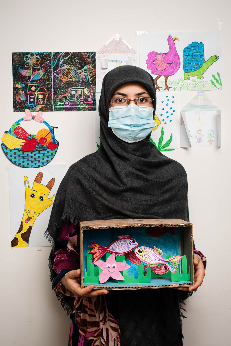Portrait of refugee Marzieh wearing glasses, a face mask and a black hijab holding a cardboard box crafted to look like a fish tank