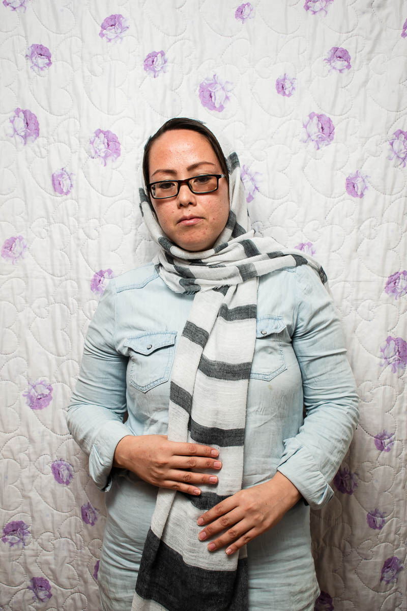 Portrait of refugee Zahra wearing glasses looking down and holding the end of her striped hijab with both her hands