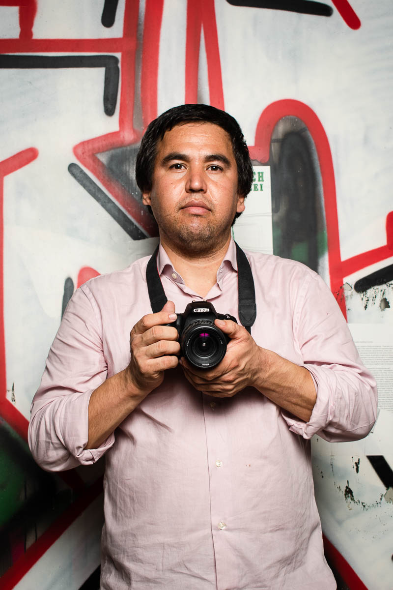 Portrait of refugee Hadi holding a camera with both his hands standing against a graffiti wall