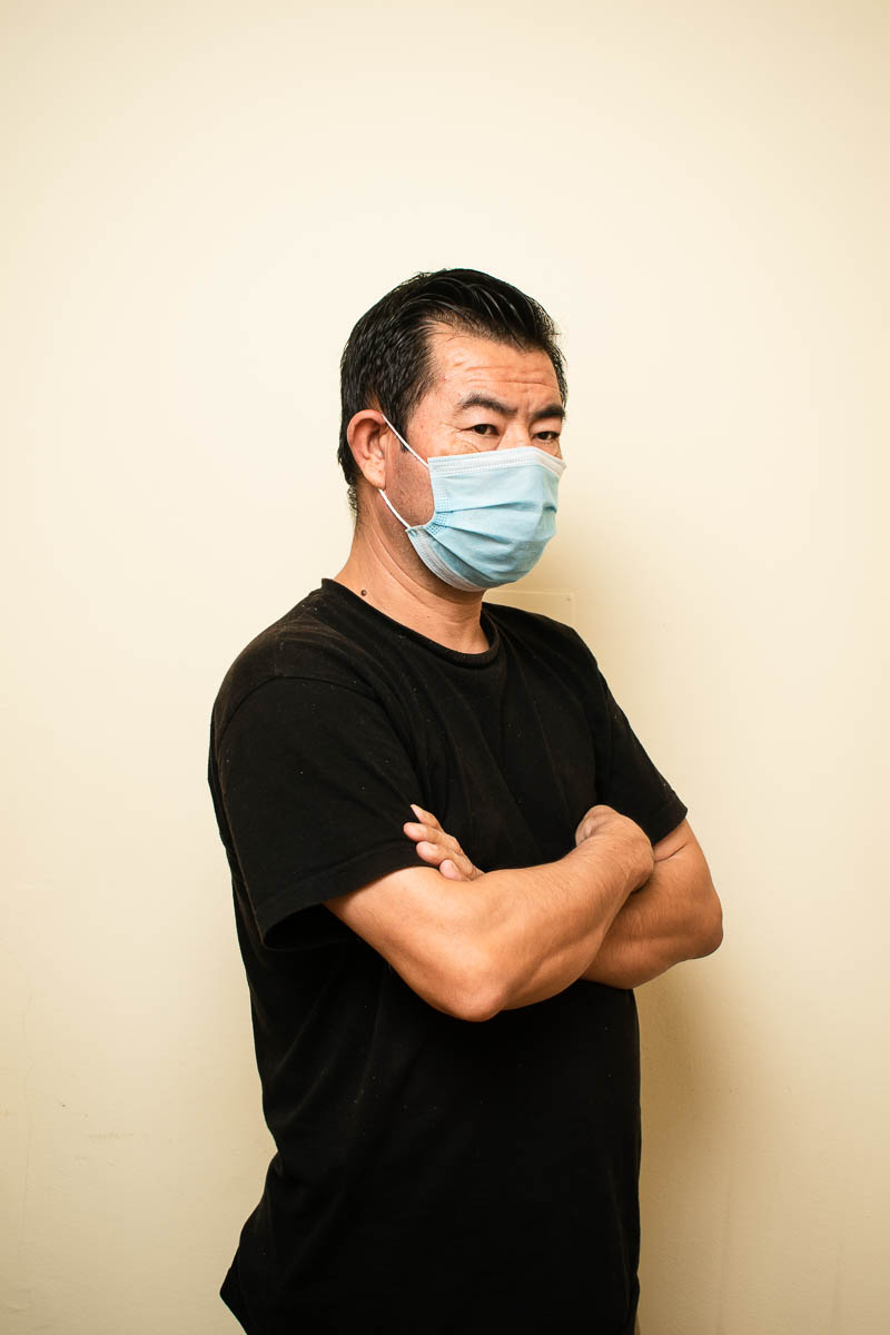 Portrait of refugee Abdul wearing a black t shirt and a face mask with his hands folded and standing sideways