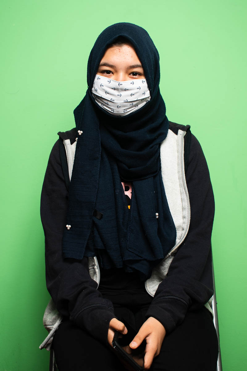 Portrait of refugee Alina wearing a black abaya and hijab with a face mask standing against a mint green wall