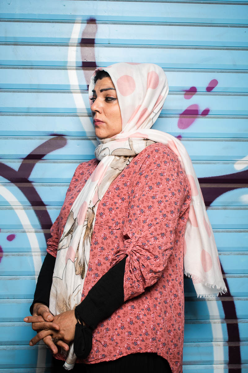 Portrait of refugee Maryam wearing a polka dotted hijab standing sideways with her hands clasped against a graffiti shutter