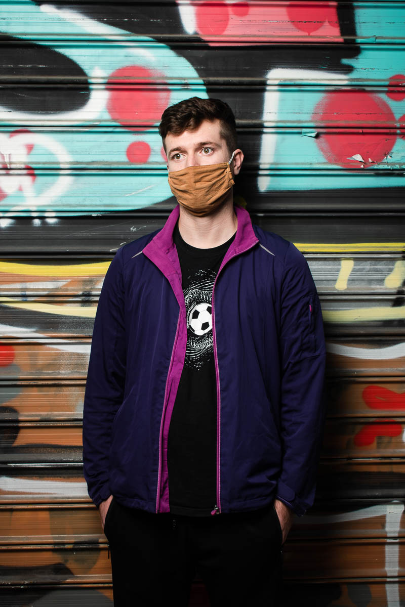 Portrait of refugee Sajad wearing a violet jacket and a brown cloth face mask with his hands in his pockets standing against a graffiti shutter
