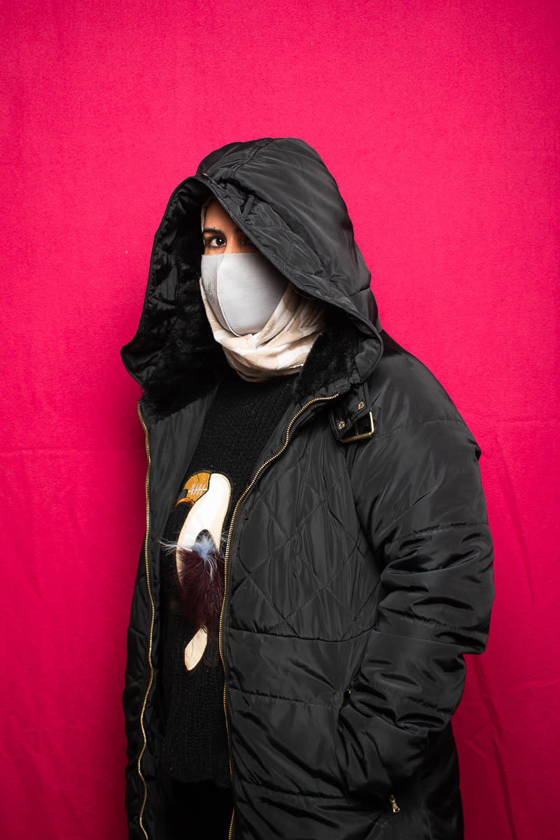 Portrait of refugee Farzaneh wearing a black hoodie and facemask to hide her face, standing against a red background