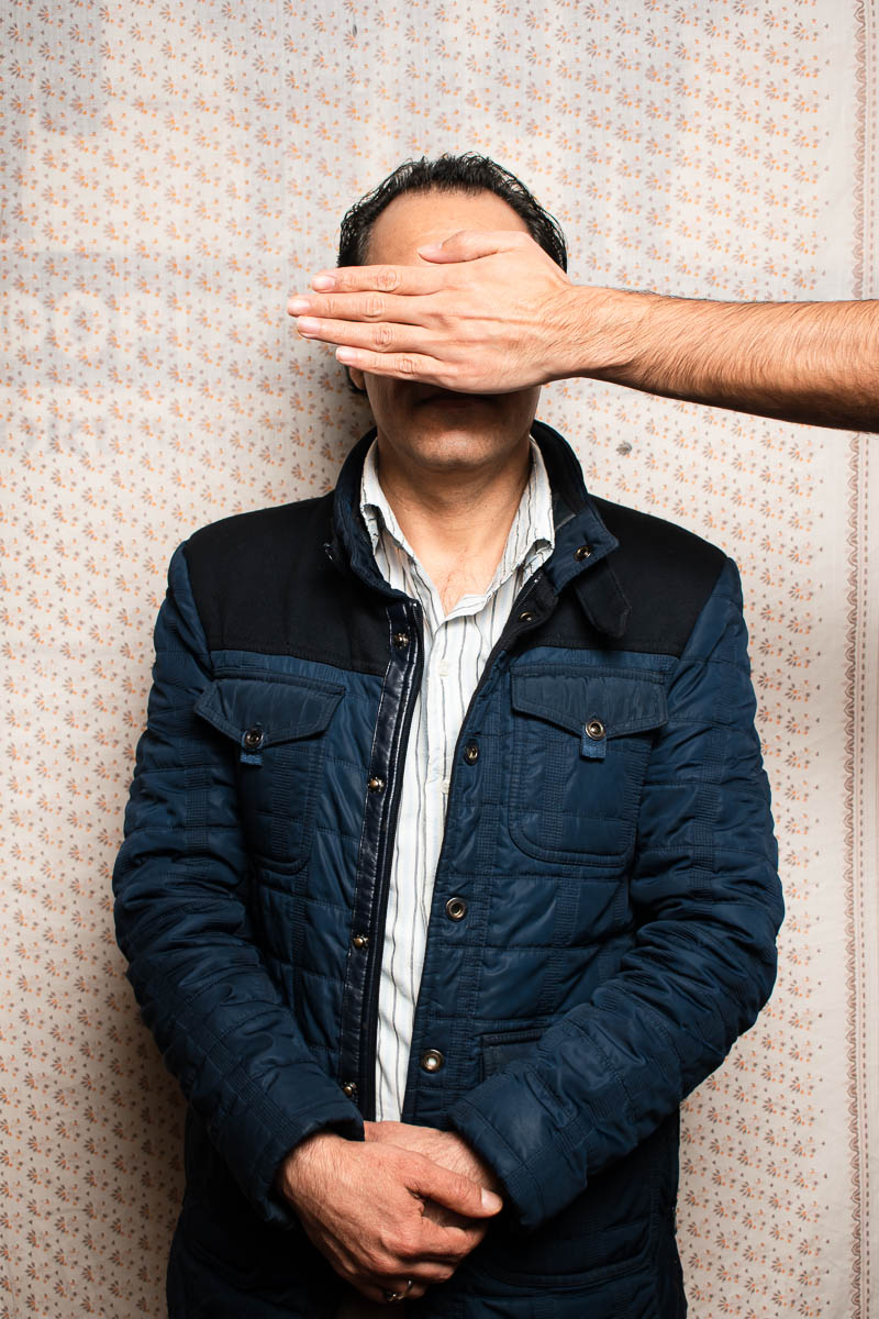 Portrait of refugee Mohamad with a hand from outside the frame covering his face while he stands with his hands clasped