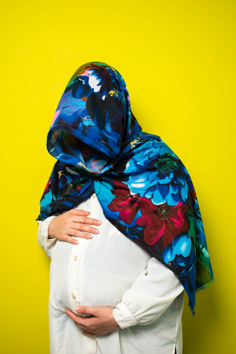 Portrait of refugee Mohadese holding her pregnant belly with a blue floral scarf covering all her face and shoulders