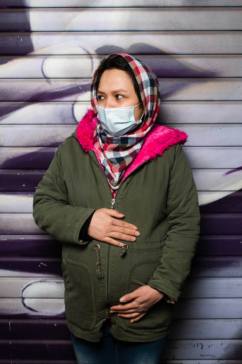 Portrait of refugee Masomeh wearing a plaid hijab and face mask holding her pregnant belly and looking to her right