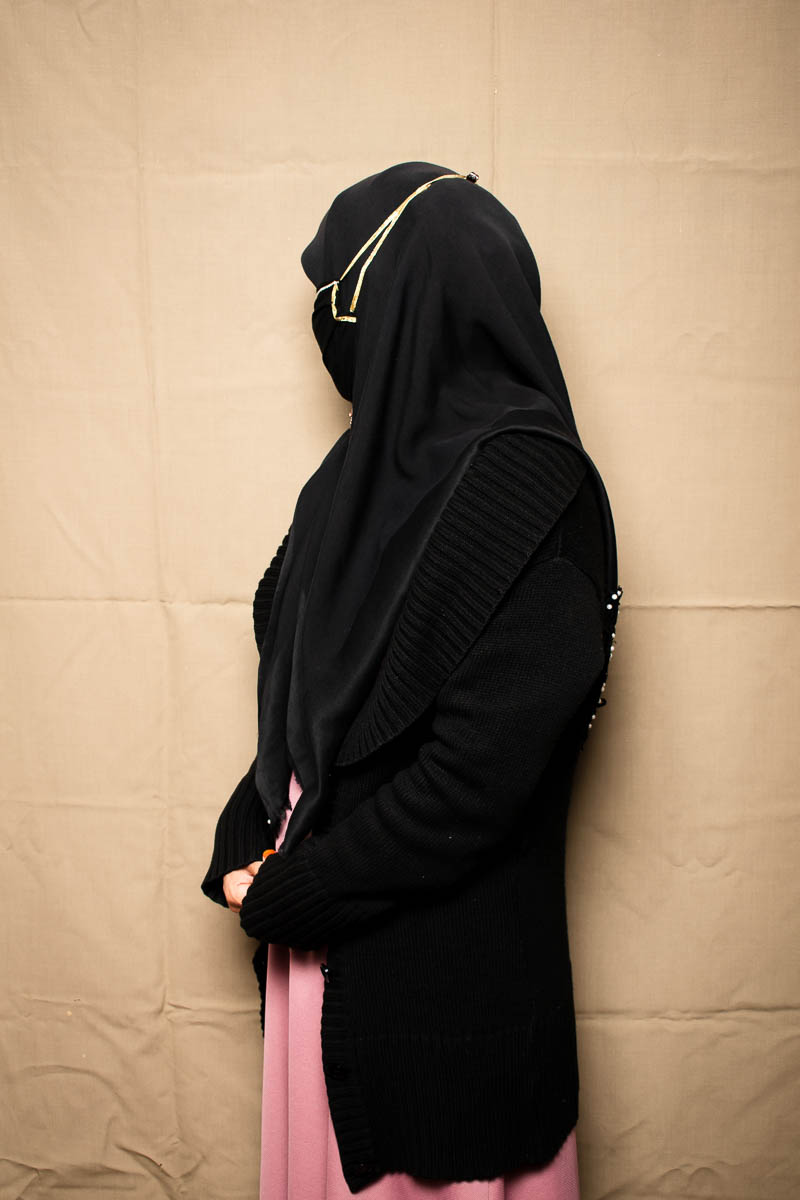 Portrait of refugee Maryam wearing a black jacket, hijab and mask with her face turned away and hidden