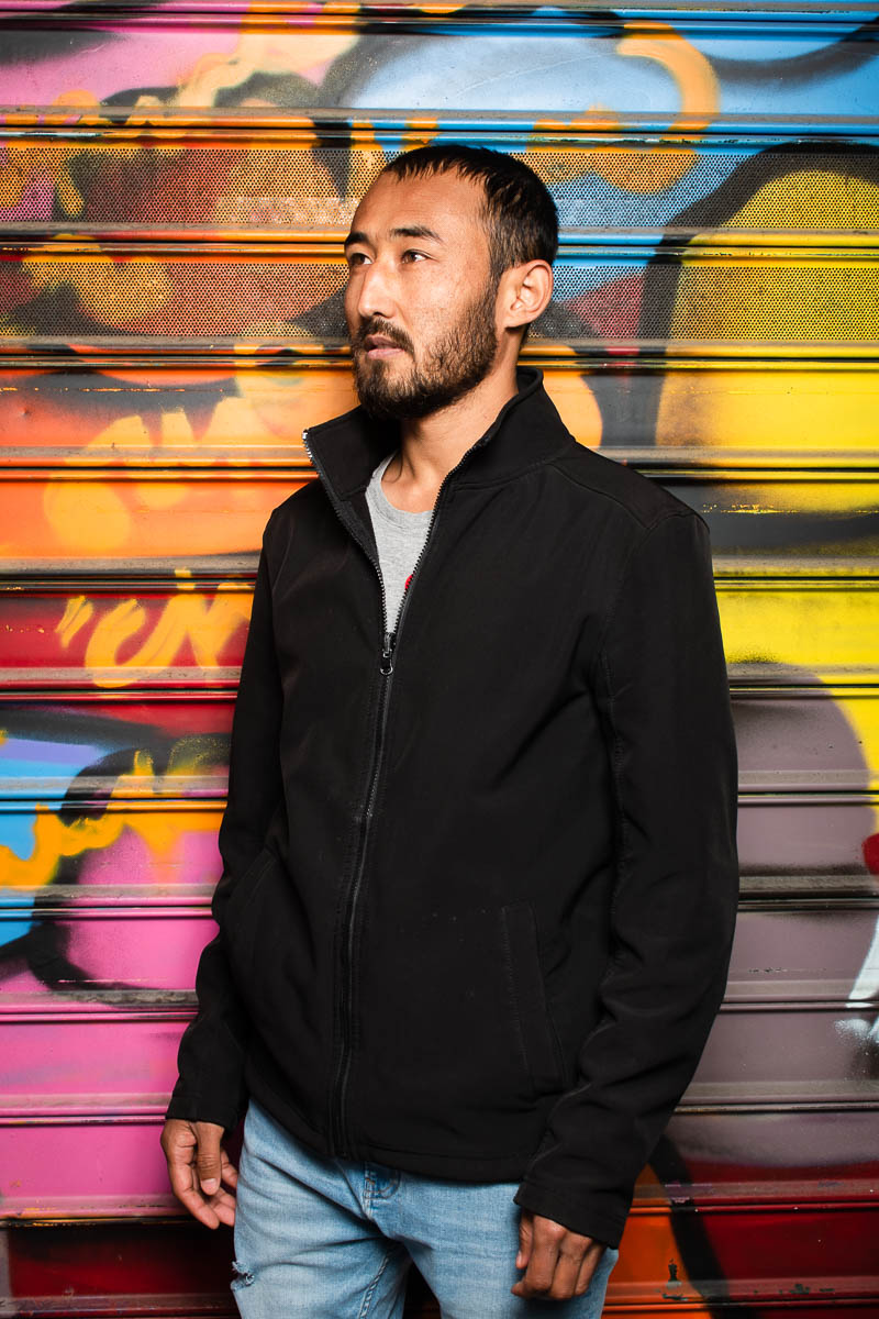 Portrait of refugee Safar wearing a black jacket turned and looking towards his right against a graffiti shutter
