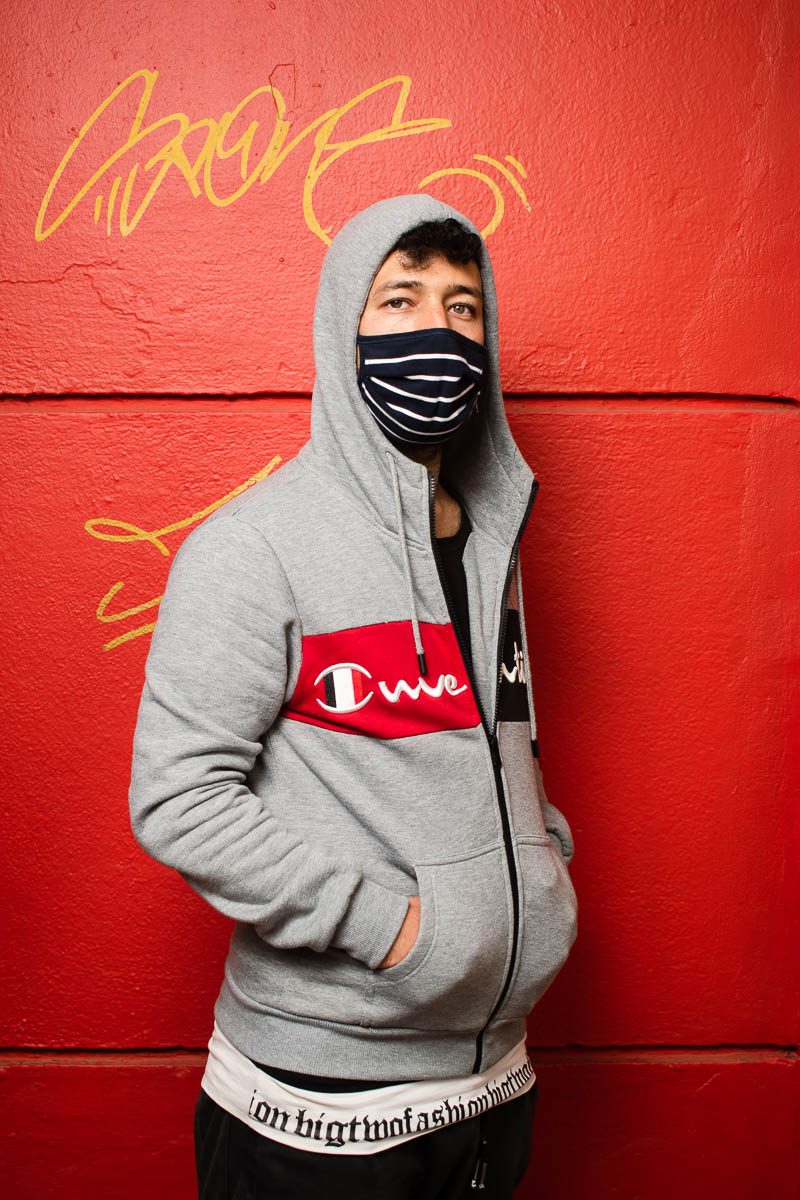 Portrait of refugee Jamal wearing a grey hoodie and black facemask with his hands in the pocket of his hoodie standing against a red wall