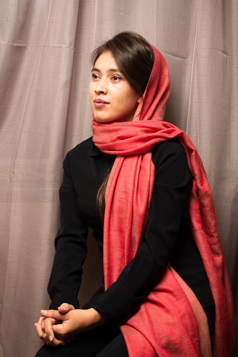 Portrait of refugee Nahid wearing a red hijab and sitting with her hands folded on her lap looking to her right
