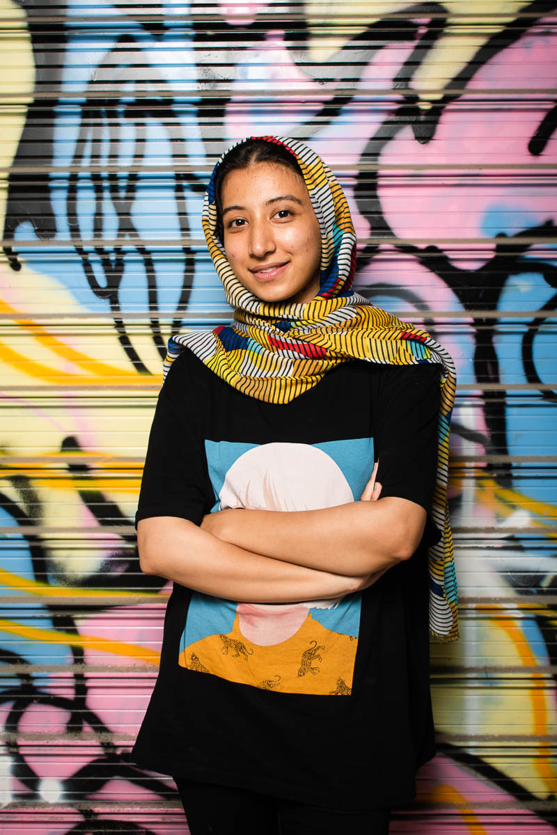 Portrait of refugee Raziyeh wearing a colorful hijab and smiling with her arms crossed standing against a graffiti container