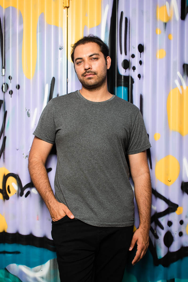 Portrait of refugee Vahid with his right hand in his pant pocket standing against a graffiti container