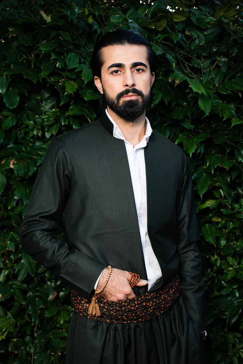 Portrait of refugee Rebin wearing traditional kurdish clothing with beads wrapped around his hand that's tucked to his belt standing against a green plat background