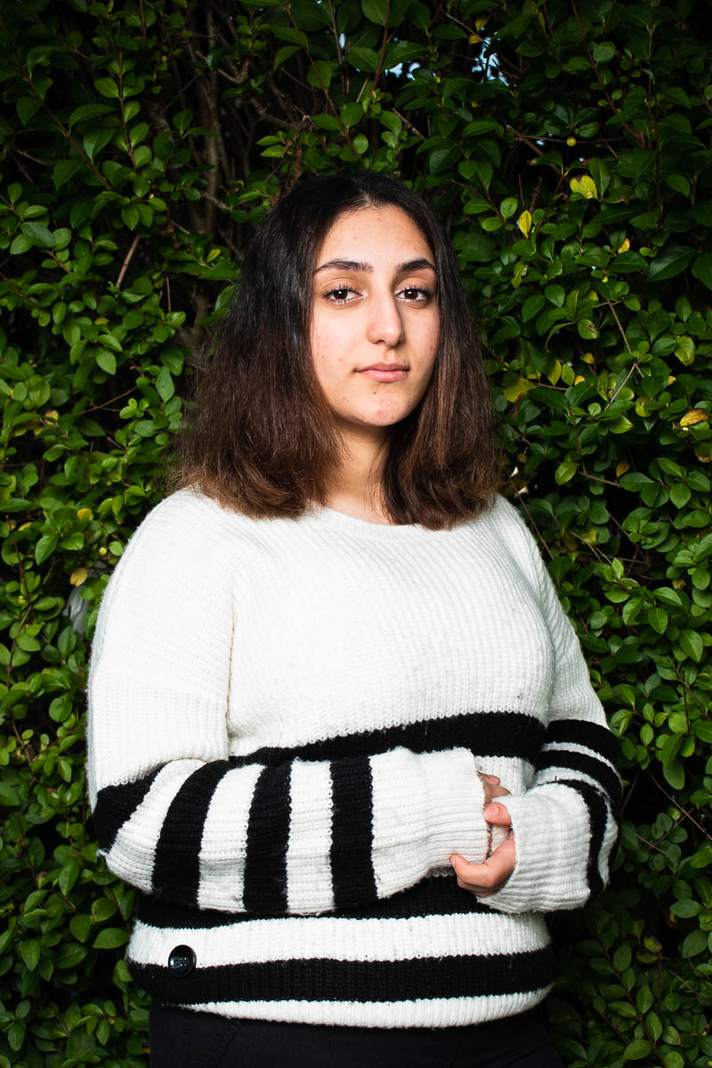 Portrait of refugee Alfat wearing a knitted sweater with her hands folded and clasped standing against a green plant background