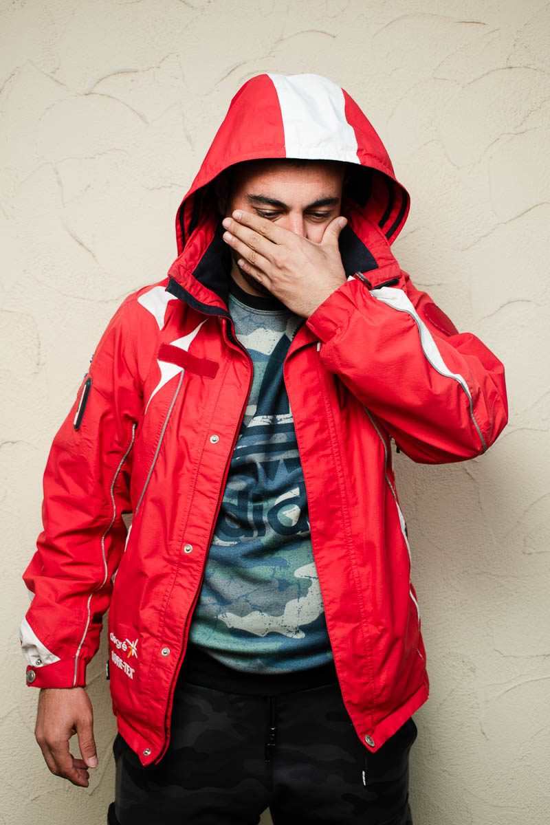 Portrait of refugee Siavash wearing a red jacket with a hoodie using his left hand to hide the lower half of his face