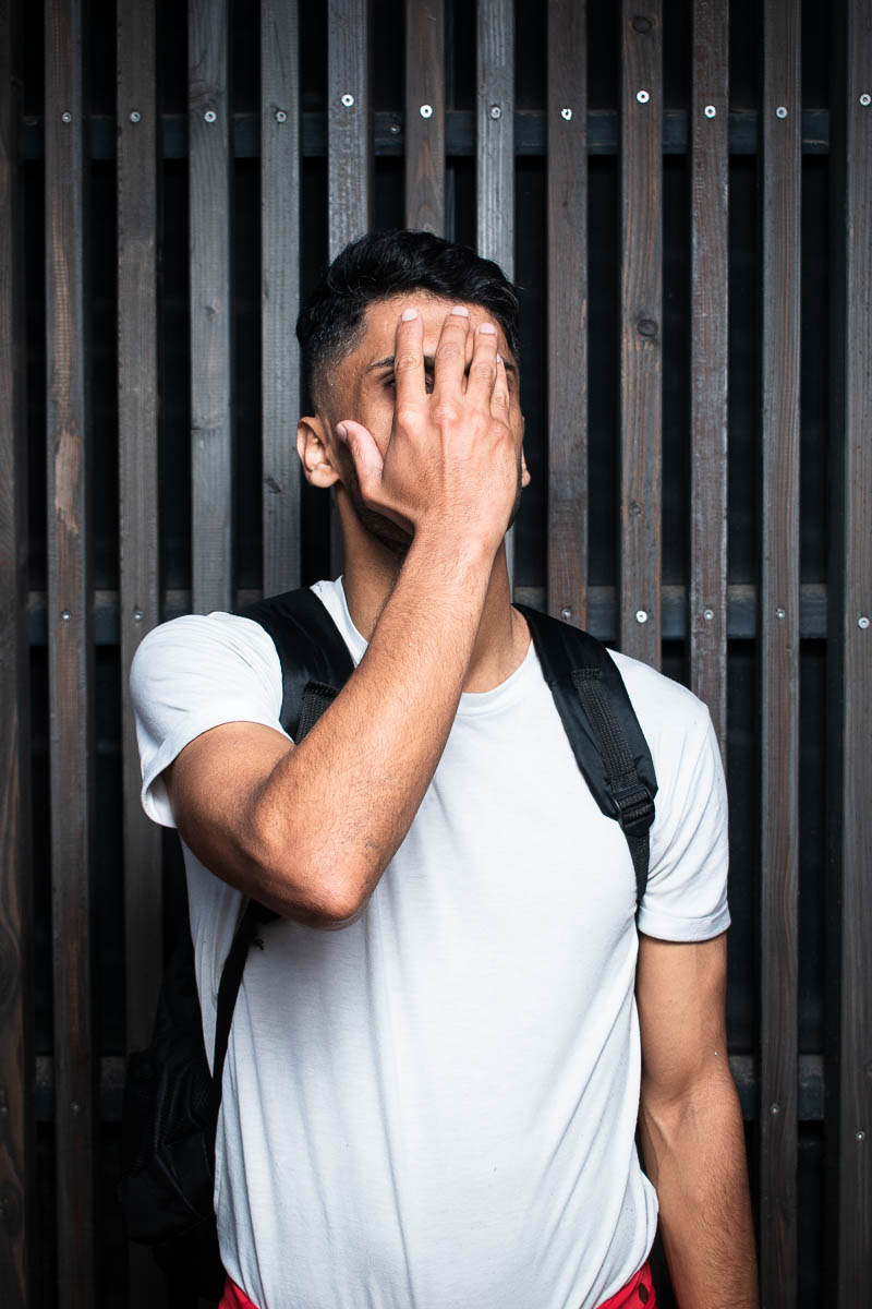 Portrait of refugee Javid wearing a white shirt with a backpack using his hand to his his face